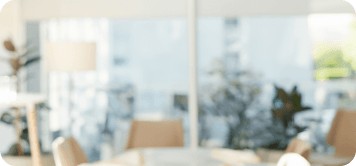 Um espaço iluminado e moderno, possivelmente um café ou uma sala de reuniões, com móveis claros e grandes janelas que deixam entrar bastante luz natural. Há plantas visíveis ao fundo.