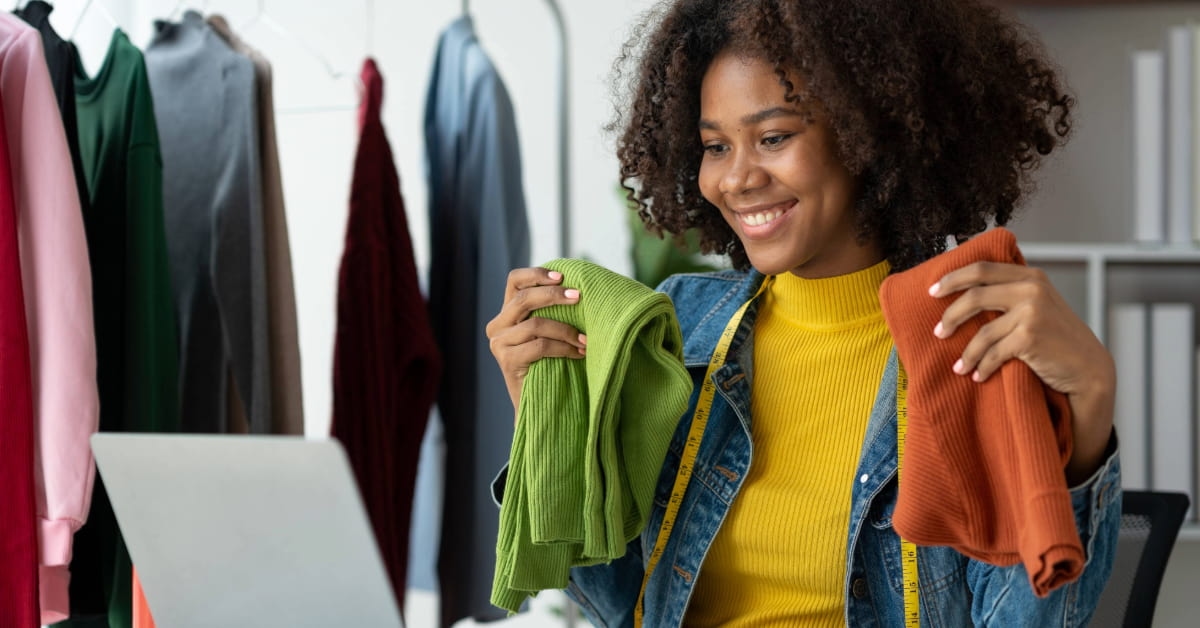 Empreendedora consultando no notebook frases para loja de roupas enquanto segura roupas do seu negócio