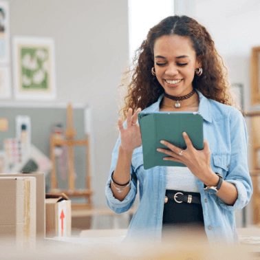 Empreendedora organizando seu cronograma de postagens Instagram em um tablet, cercada por caixas e materiais de escritório.
