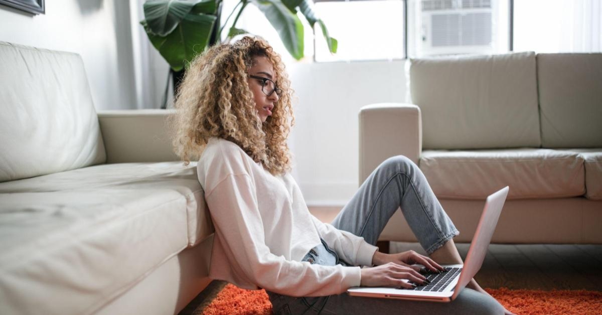 A imagem mostra uma mulher preta, de cabelos encaracolados com mechas loiras. Ela está sentada no chão, apoiada em um sofá, enquanto escreve em um notebook. Uma alusão ao momento de pesquisa sobre como criar um produto de impacto.