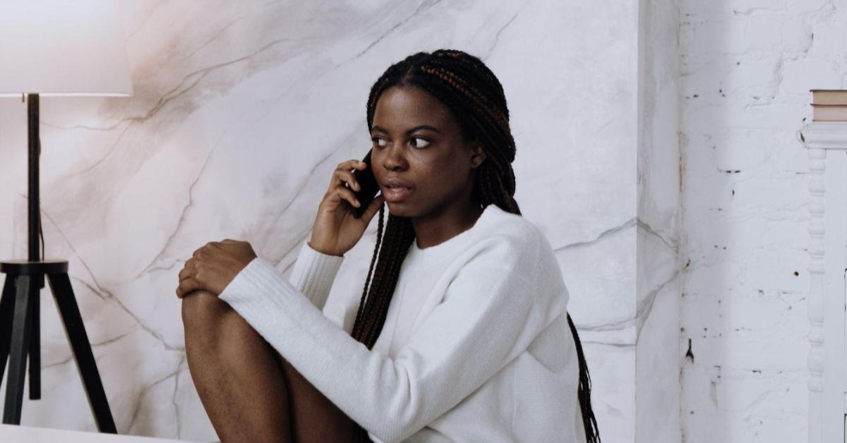 A imagem mostra uma mulher preta, de cabelos trançados, vestindo uma camisa branca de mangas compridas. Ela está sentada em uma cadeira, de pernas cruzadas, enquanto fala no celular. Uma alusão a como vender no whatsapp pode ser uma estratégia eficaz para os pequenos negócios.