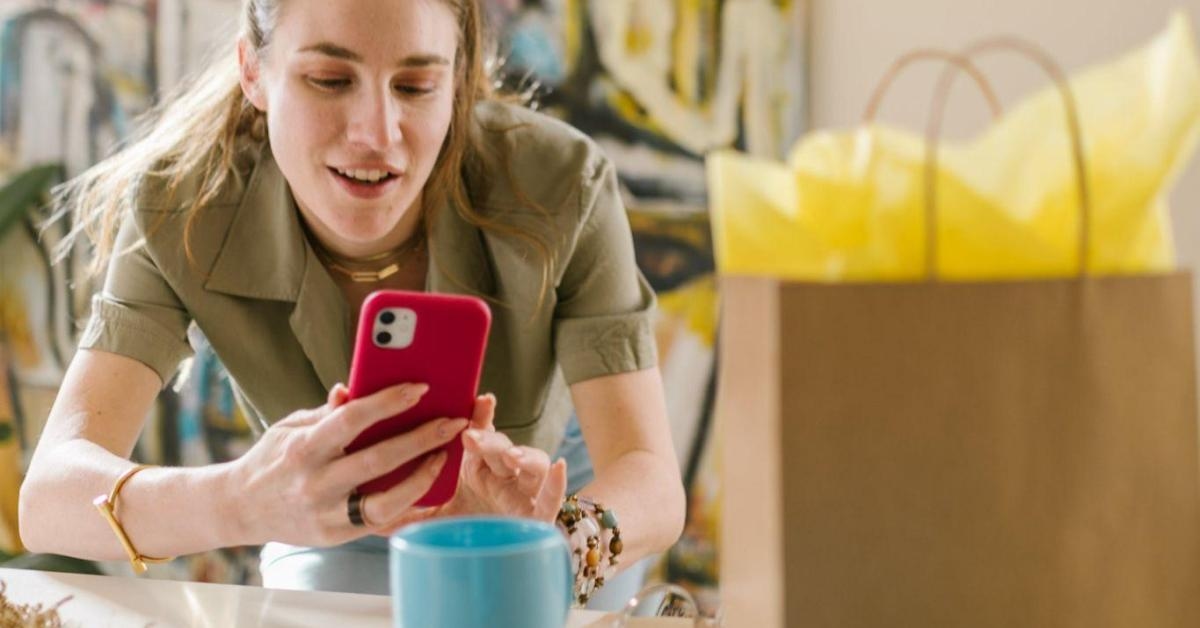 A imagem mostra uma mulher branca, loira, que veste uma camisa polo verde musgo, segurando um celular vermelho. Ela está focada em tirar fotos de uma caneca azul, que está sobre uma mesa, ao lado de uma sacola de presente. A imagem remete a importância das fotos de produto no e-commerce, uma das maneiras que ajuda lojas a aparecer no google