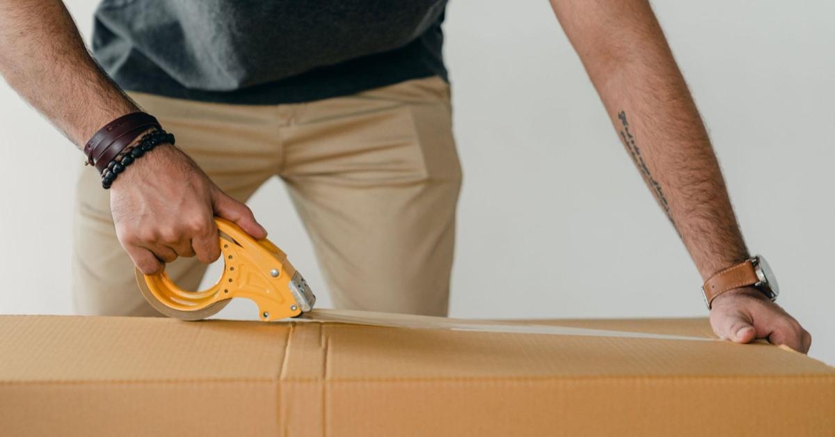 A imagem mostra um homem, de calça bege e camisa polo azul escuta, fechando um pacote de papelão com fita adesiva. Na imagem o hosto do homem não parece, apenas seus braços e parte do tronco. Em seus braços há pulseiras, e no bolso da calça um celular. Aqui, temos uma idea de uma das etapas necessárias para enviar um pacote via Sedex.
