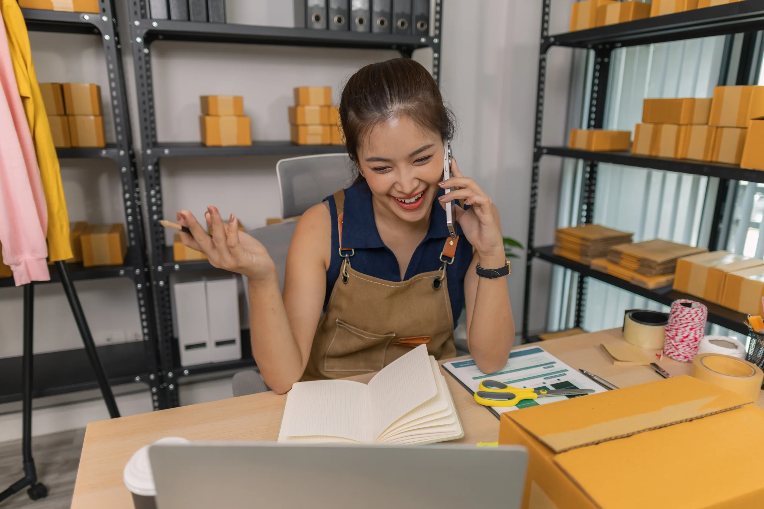 Mulher empreendedora conversando pelo celular enquanto organiza pedidos e embalagens para divulgação de produtos e vendas online.